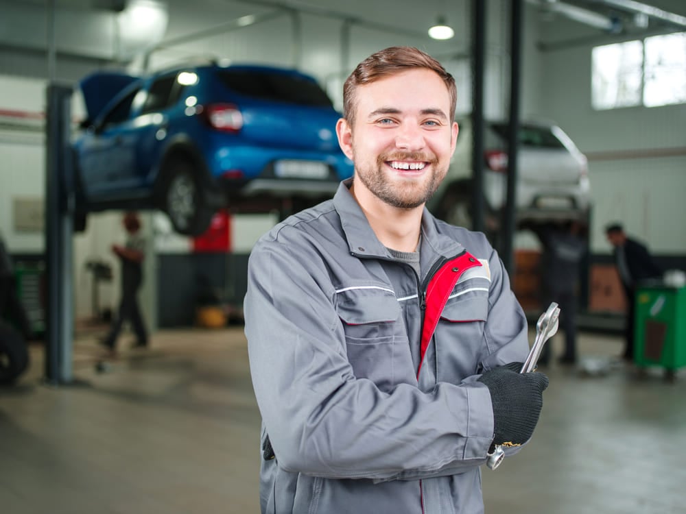 Smiling repair mechanic with an auto repair garage and cars behind him